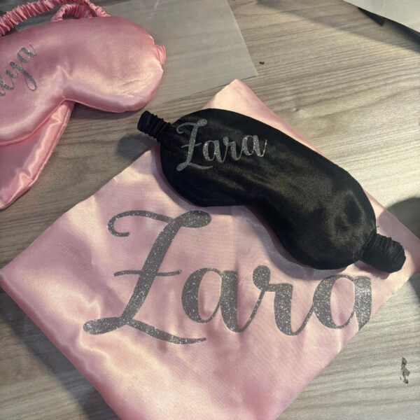 Black and pink sleep mask with 'Lara' printed on a pink satin bag on a wooden surface
