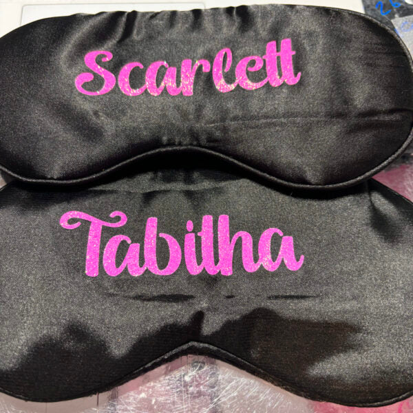 Black satin sleep mask with pink embroidered names on a fabric background