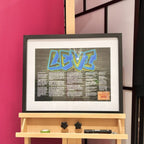Framed speech gift artwork with 'LEVI' in colourful letters on a wooden shelf against a pink wall with no acrylic