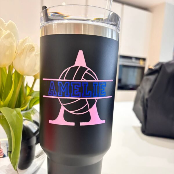 Black stanley-style cup with pink and blue design and 'AMELIE' text, placed on a surface with flowers and a bag in the background.