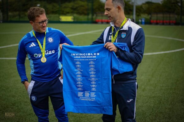 Two men on a sports field holding a blue t-shirt with 'Invincibles' printed on it.