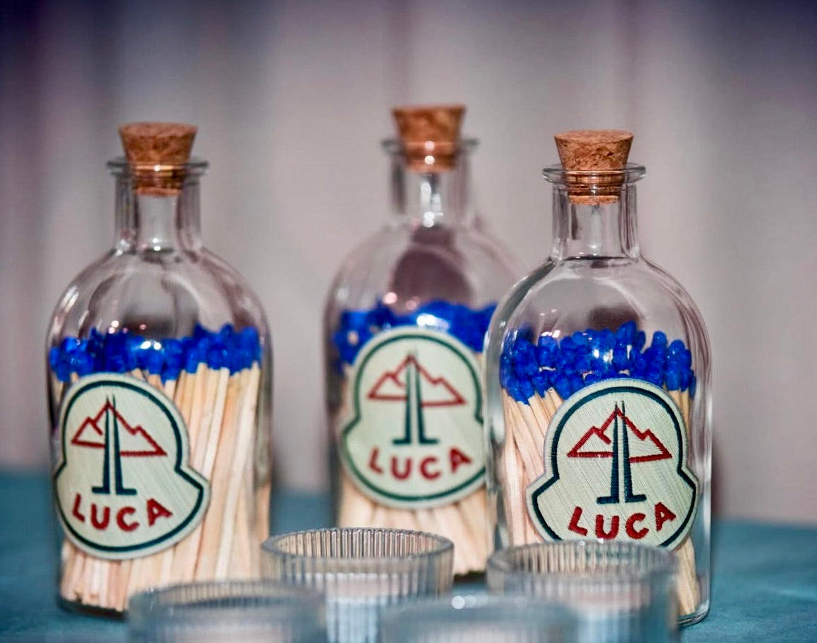 Three glass match stick bottles with cork lids containing matches labelled 'LUCA' on a blurred background.