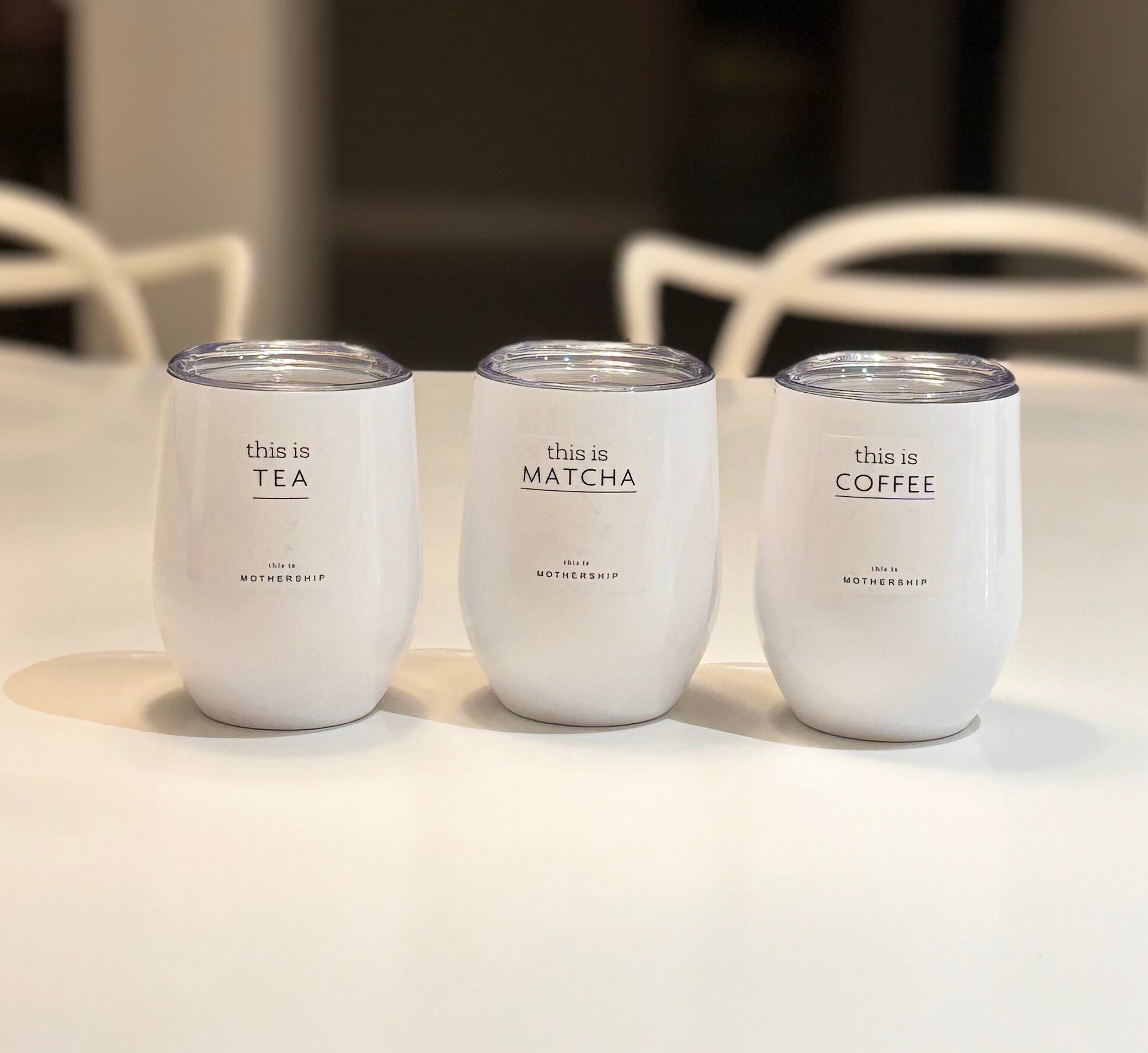 Three white travel cups labelled 'this is TEA', 'this is MATCHA', and 'this is COFFEE' on a light surface.