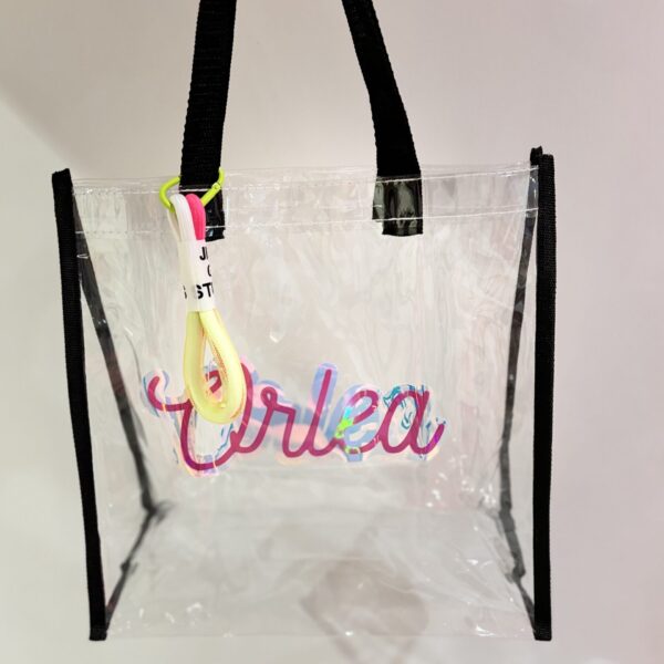 Clear tote bag with colourful text and black straps on a light grey background