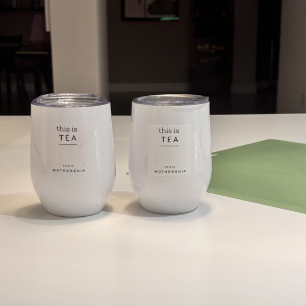 Two white travel cups labelled 'this is TEA' on a blurred light surface.