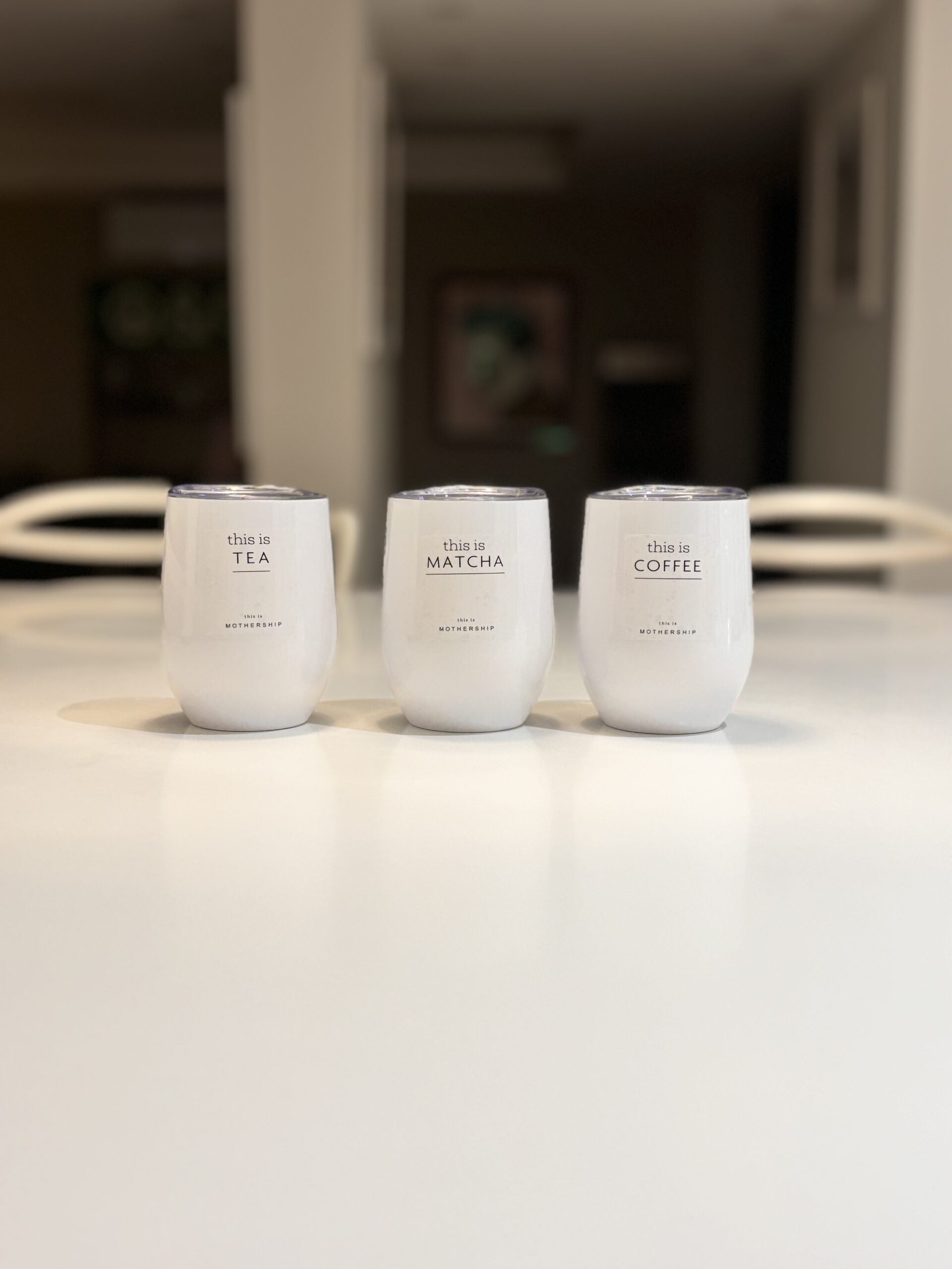 Three white travel cups labelled 'this is TEA', 'this is MATCHA', and 'this is COFFEE' on a blurred light surface.