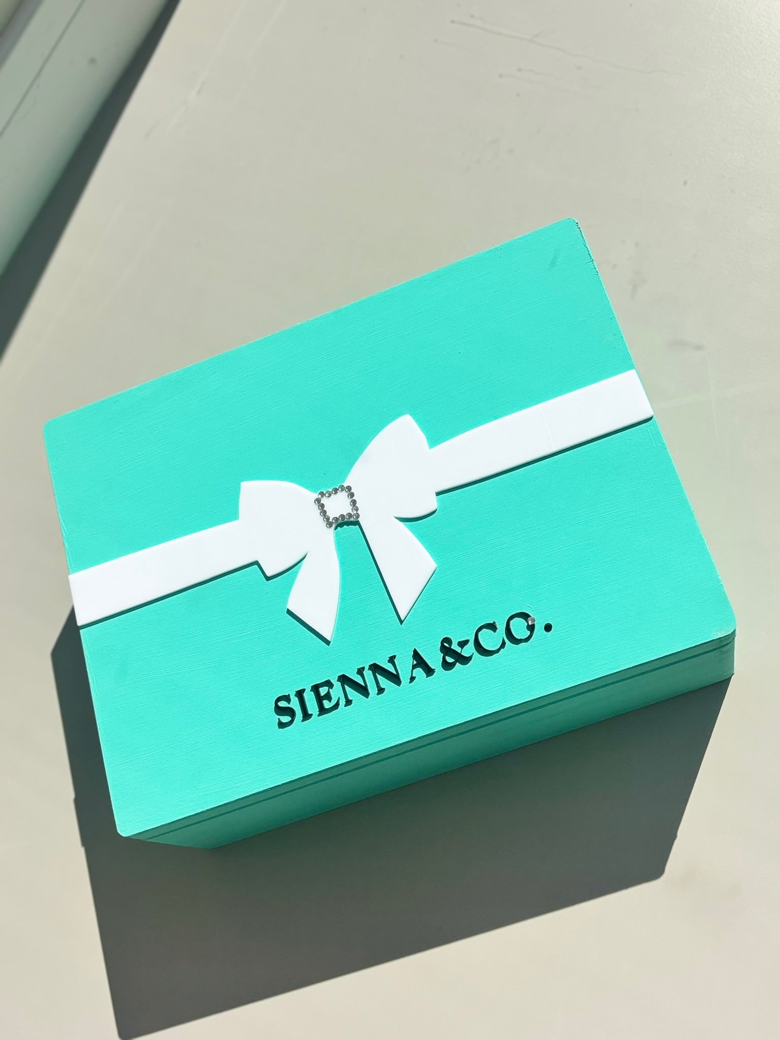 Turquoise keepsake box with white ribbon and 'SIENNA & CO.' branding on a light grey background