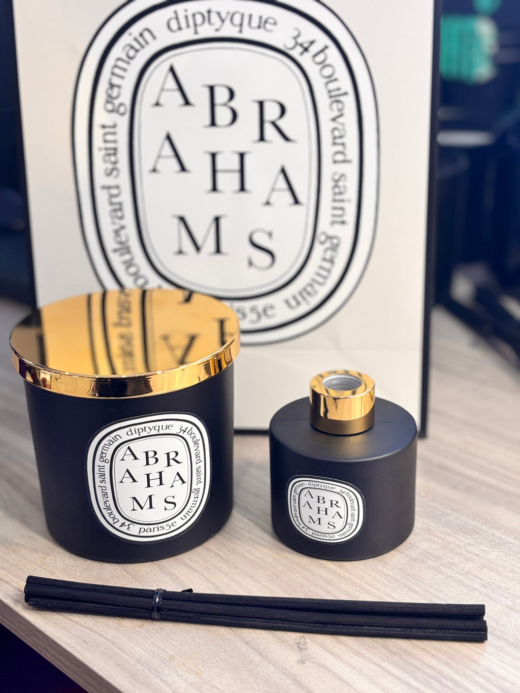 Two 'Abrahams' candle and diffuser set with gold tops on a wooden surface, and branded bag in the background.