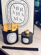 Two 'Abrahams' candle and diffuser set with gold tops on a wooden surface, and branded bag in the background.