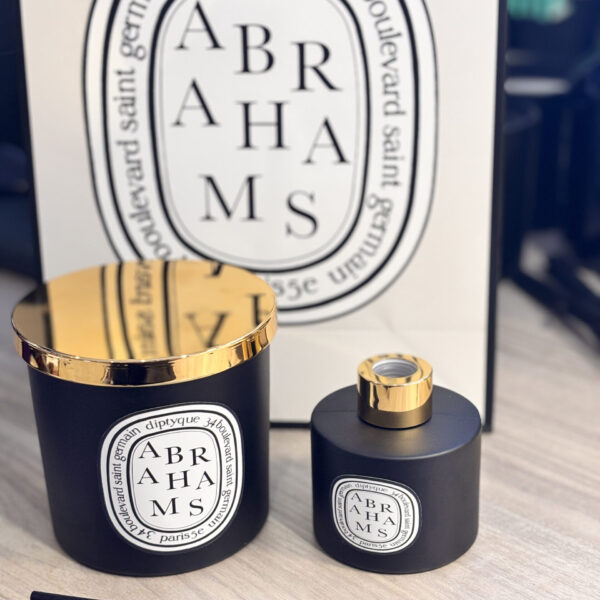 Two black candle and diffuser set with gold caps and 'ABRAHAMS' branding in front of a Diptyque-style sign.