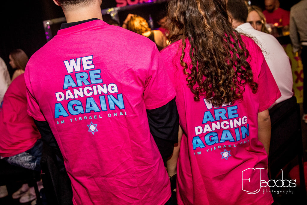 Two people wearing pink event t-shirts with 'We Are Dancing Again' text at a social event.