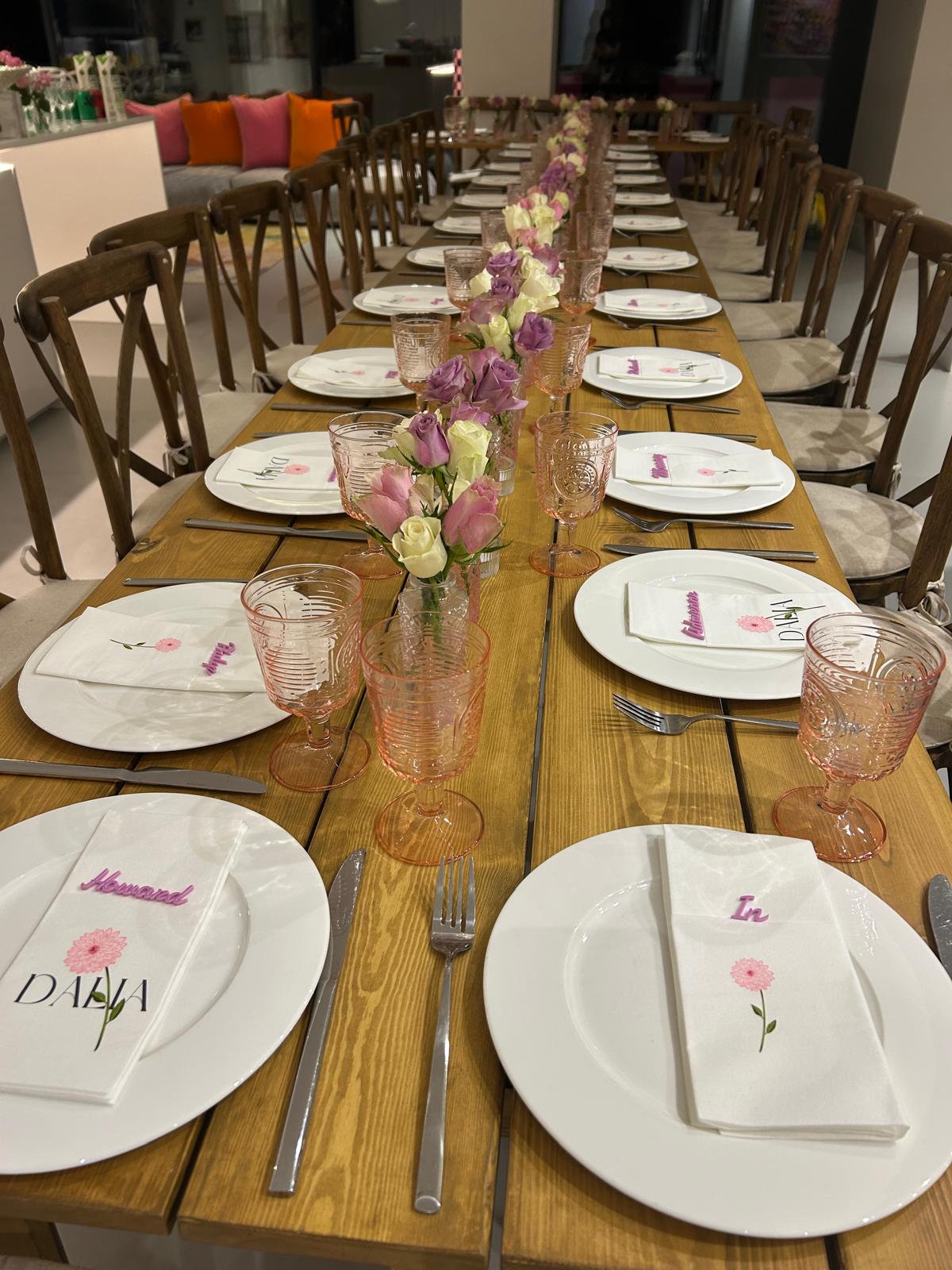 Long wooden dining table set for a formal dinner with acrylic place name settings, glasses, and floral centre pieces.