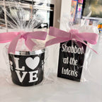 Two gift candles wrapped in cellophane with pink ribbons, one with 'LOVE' text and the other with 'Shabbat at the Lutons' text.
