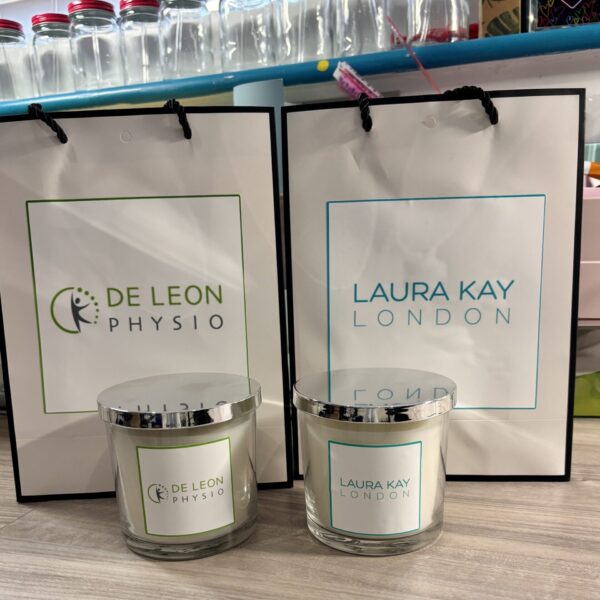 Two candles with 'De Leon Physio' and 'Laura Kay London' branding in front of branded bags.