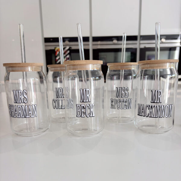 Set of clear cold cups with wooden lids and names printed on them, placed on a white surface.