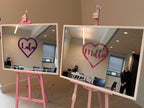 Two classic mirrors on pink stands with 'Lulu' and 'Mollie' written in hearts, in an office setting.
