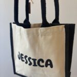 Beige personalised tote bag with 'JESSICA' printed on it, held by a hand against a neutral background.