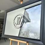 Grey-framed bespoke mirror with 'JK' logo on a wooden stand indoors