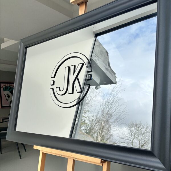 Grey-framed bespoke mirror with 'JK' logo on a wooden stand indoors