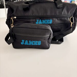 Black duffel bag and washbag and small pouch with 'JAMES' printed on them, placed on a light surface.