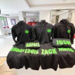 Black hoodies with green names on mannequins in a room.