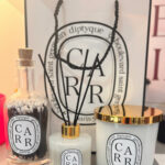 Diptyque style dffuser, candle, and match bottle with decorative sticks against a Diptyque-style  branded background.
