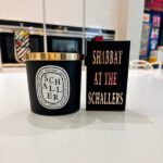 Gift set of a black Schaller candle with gold lid on a white surface, next to a black match box sleeve with 'Shabbat at the Schallers' text.