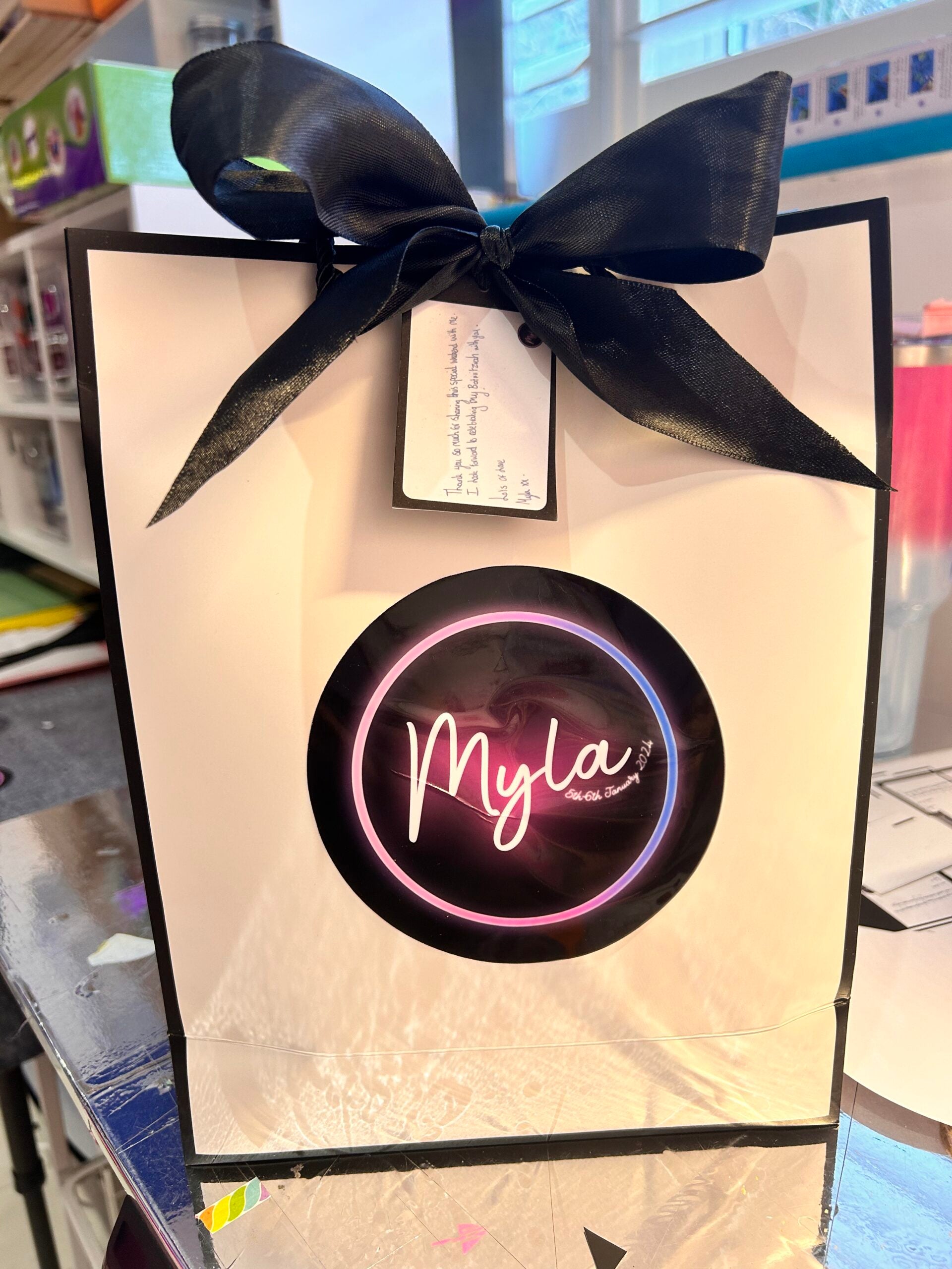 Gift bag with a black bow and 'Myla' logo on a blurred indoor background