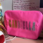 Pink washbag with 'GABRIELLA' in glittery letters on a desk with papers in the background