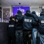 Group of people wearing hoodies with names on the back, gathered around a screen displaying a logo.