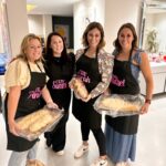 Four women in a kitchen holding bread, wearing personalised aprons with names.