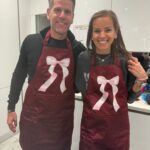 Two people wearing personalised maroon aprons with pink bows in a kitchen.