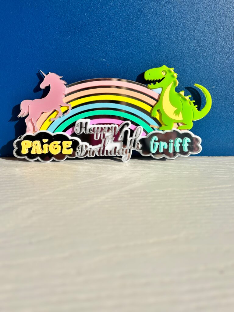 Colorful birthday acrylic cake topper with unicorn, rainbow, and dinosaur on a blue background