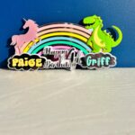 Colorful birthday acrylic cake topper with unicorn, rainbow, and dinosaur on a blue background