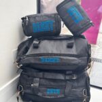 Black washbags and duffel bags with names embroidered on them, including 'DANNE' and 'SETH', against a blurred indoor background.
