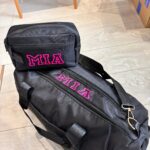 Black duffel bag and washbag with 'MIA' printed in pink on a tiled floor.