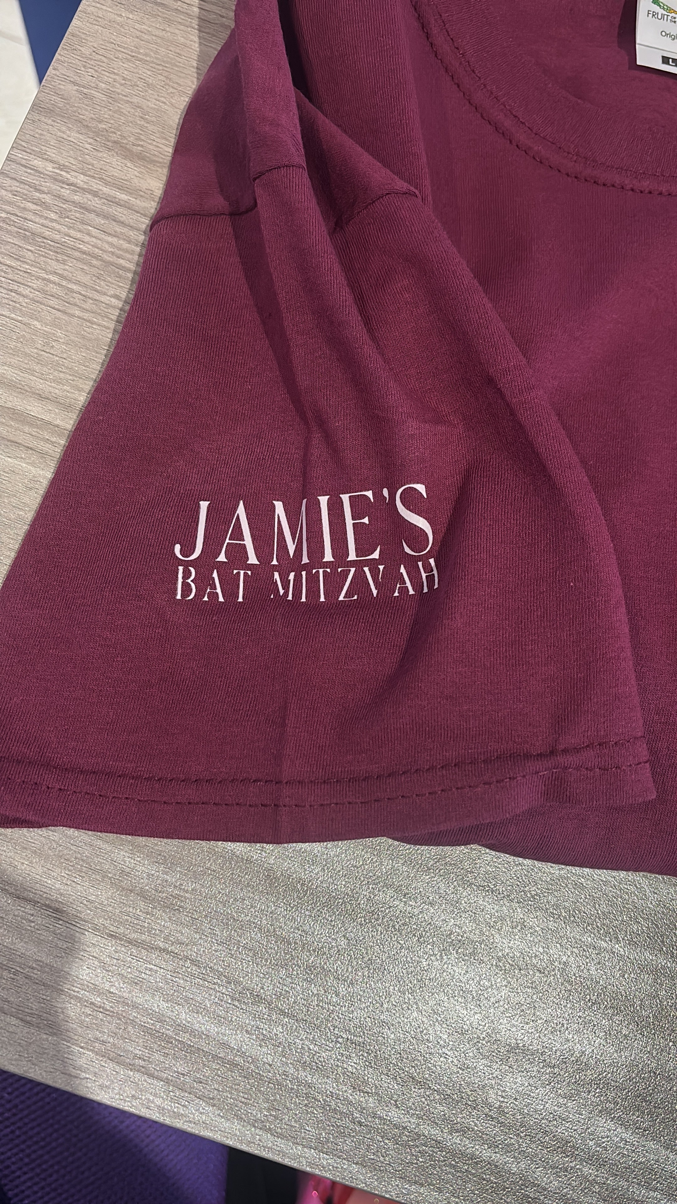 Maroon event t-shirt with 'JAMIE'S BAT MITZVAH' text on a textured surface