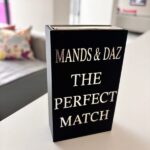 Black match box sleeve with 'The Perfect Match' by MANDS & DAZ on a table in a room.