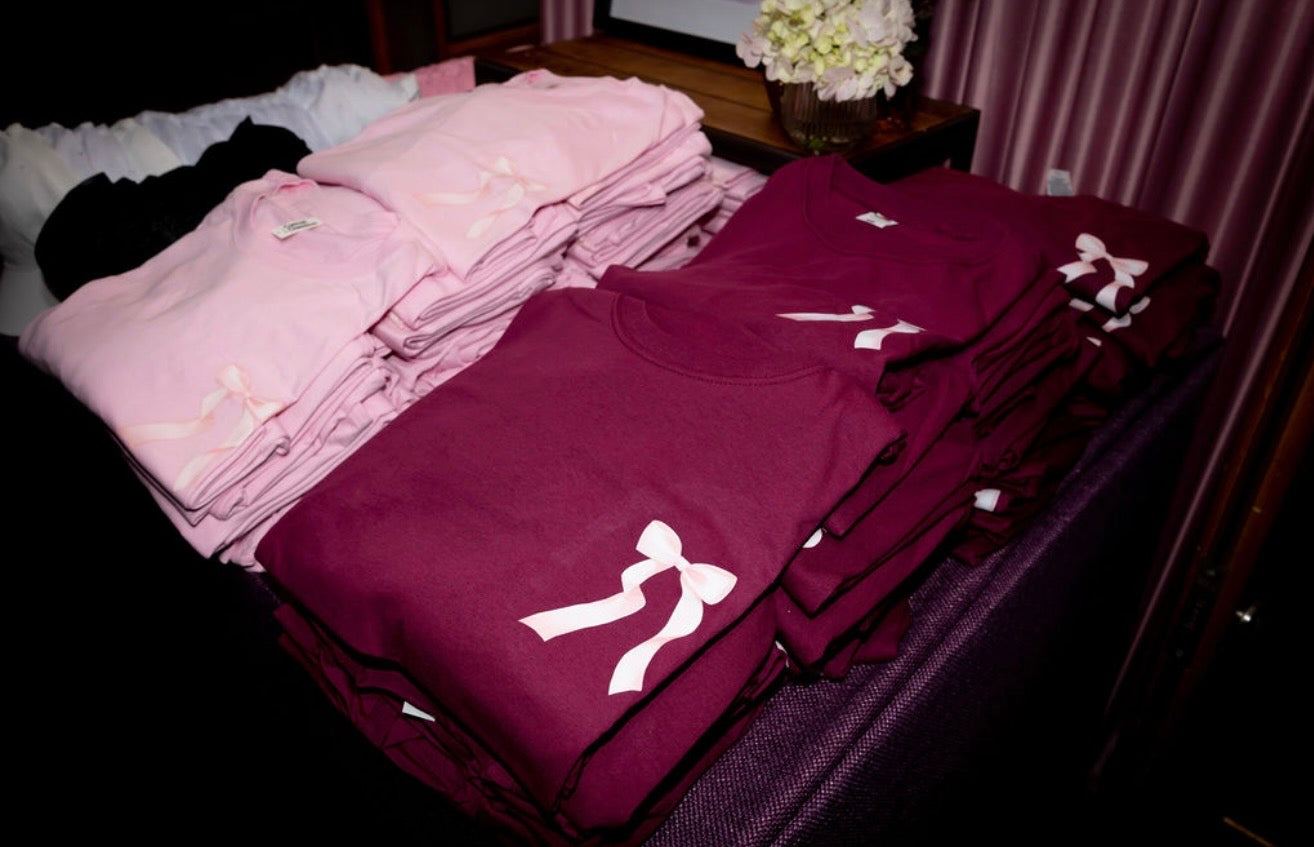 Stack of maroon and pink event t-shirts with white designs on a dark surface.
