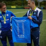 Two men on a sports field holding a blue t-shirt with 'Invincibles' printed on it.