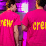 Two people wearing pink 'crew' personalised t-shirts in a dimly lit room with purple lighting.