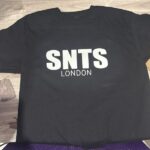 Black t-shirt with 'SNTS LONDON' text on a wooden surface