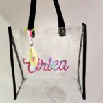 Clear tote bag with colourful text and black straps on a light grey background