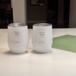 Two white travel cups labelled 'this is TEA' on a blurred light surface.