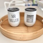 Two white travel cups with personalised text on a wooden tray.
