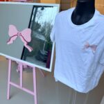 White event t-shirt with pink bow design on a mannequin, displayed next to a mirror reflection.