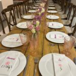 Long wooden dining table set for a formal dinner with acrylic place name settings, glasses, and floral centre pieces.