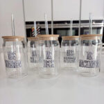 Set of clear cold cups with wooden lids and names printed on them, placed on a white surface.