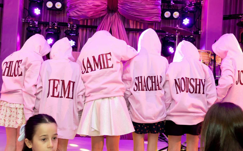 Group_of_people_wearing_pink_hoodies_with_names_printed_on_them_standing_in_front_of_a_stage_with_purple_lighting_taken_for_Mirrored_Memories