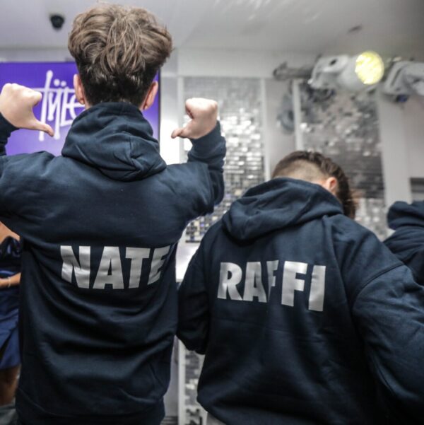 Two_people_wearing_dark_hoodies_with_NATE_and_RAFFI_printed_on_the_back_in_a_room_with_a_purple_screen_and_lights.